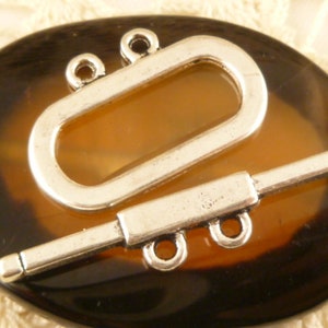 Può includere: Chiusura a scatto in metallo argentato con una barra rettangolare e un anello ovale. La chiusura è su uno sfondo marrone.