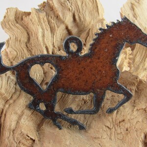 May include: A brown metal horse pendant with a heart-shaped cutout. The pendant has black outlining and a loop for hanging. The horse is depicted in a running pose, set against a textured wooden background. The pendant is likely for a necklace.