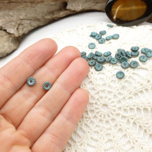 May include: Small, turquoise-colored metal beads with a floral design. The beads are scattered on a white lace surface, with a few held in a hand. The beads have a distressed, vintage appearance.
