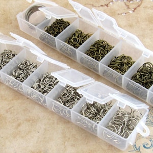 May include: Two clear plastic organiser boxes filled with various sizes and colours of jump rings. The jump rings are silver, bronze, and gunmetal. The boxes have individual compartments with hinged lids.