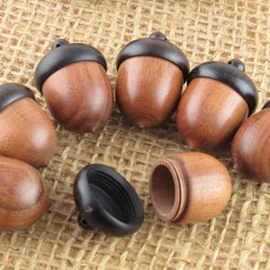 May include: A selection of wooden acorn pendants with dark brown caps. One acorn is open, showing the hollow inside. The acorns are light brown and are on a textured, tan surface.