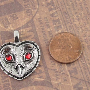 Peut inclure: Pendentif en argent en forme de c&oelig;ur avec un motif de hibou. Le hibou a des yeux rouges et un visage d&eacute;taill&eacute;. Le pendentif est &agrave; peu pr&egrave;s de la taille d'une pi&egrave;ce de monnaie d'un centime d'euro.