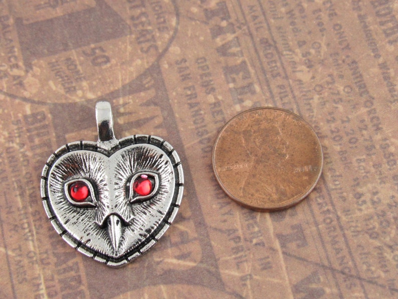 Peut inclure: Pendentif en argent en forme de c&oelig;ur avec un motif de hibou. Le hibou a des yeux rouges et un visage d&eacute;taill&eacute;. Le pendentif est &agrave; peu pr&egrave;s de la taille d'une pi&egrave;ce de monnaie d'un centime d'euro.
