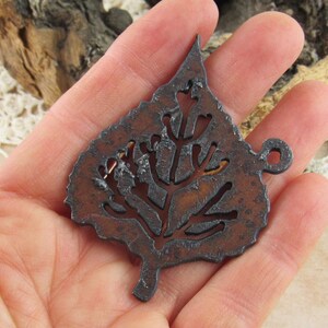 May include: A rustic, brown metal leaf pendant with intricate cut-out details. The pendant has a loop for hanging and is held in a hand. The leaf design features a detailed vein pattern and a textured surface, perfect for jewelry making or crafting.