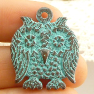 May include: A turquoise blue metal owl charm with a textured surface. The owl has large eyes, a beak, and wings. The charm has a loop at the top for attaching to a necklace or bracelet.