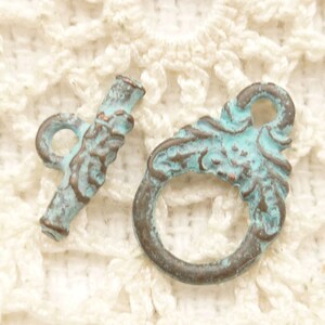 May include: Two antique-style toggle clasps with a distressed finish. The clasps feature a turquoise and bronze colour scheme, with intricate floral detailing. The toggle is a bar shape, and the other clasp is a ring shape.