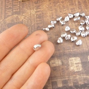 May include: A pile of silver metal beads in the shape of a teardrop with a hole in the top. The beads are scattered on a brown surface.