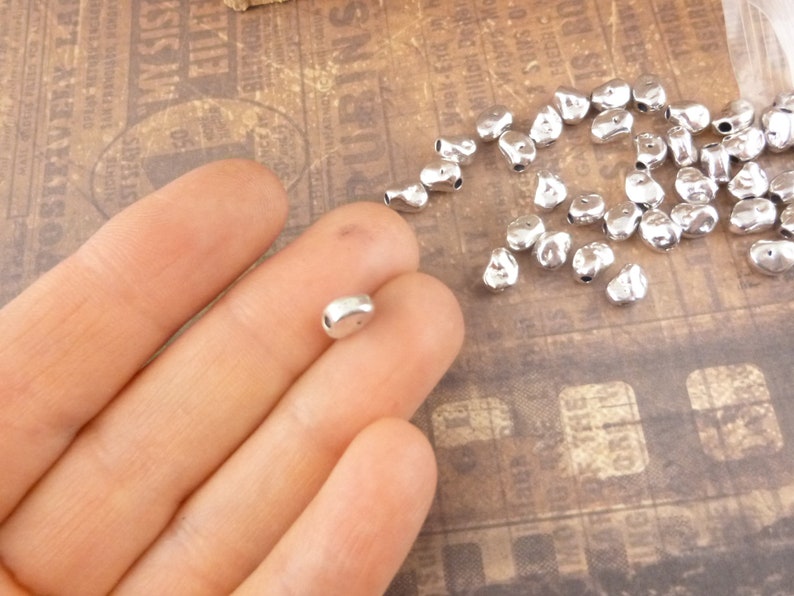 May include: A pile of silver metal beads in the shape of a teardrop with a hole in the top. The beads are scattered on a brown surface.