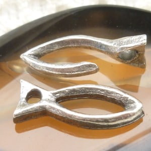 Puede incluir: Dos colgantes plateados con forma de pez, cada uno con un agujero en la parte superior. Los colgantes tienen una superficie texturizada y están colocados sobre un fondo marrón claro y negro. Estos colgantes se pueden utilizar para hacer joyas u otros proyectos de artesanía.