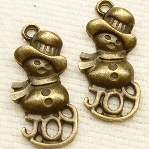 Puede incluir: Dos abalorios de muñeco de nieve de metal dorado antiguo con la palabra "JOY" escrita en la parte inferior de cada abalorio.