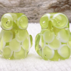 Puede incluir: Dos pequeñas figuras de búho de cristal. Los búhos son de color verde claro con detalles blancos. El cristal es translúcido y los búhos tienen ojos grandes y redondos y pequeñas alas.