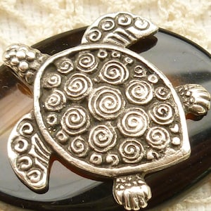 May include: A silver turtle pendant with a swirling pattern on its shell, set on a black and brown agate stone.