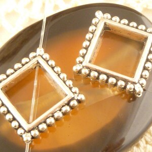 May include: Two silver-toned metal picture frame pendants with a square opening and a beaded border. The pendants are on a brown background.