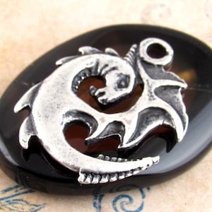 May include: A silver-colored pendant in the shape of a stylized dragon and anchor, set on a dark, oval-shaped stone. The pendant has intricate details and is likely made of metal. The stone has a polished, glossy finish.