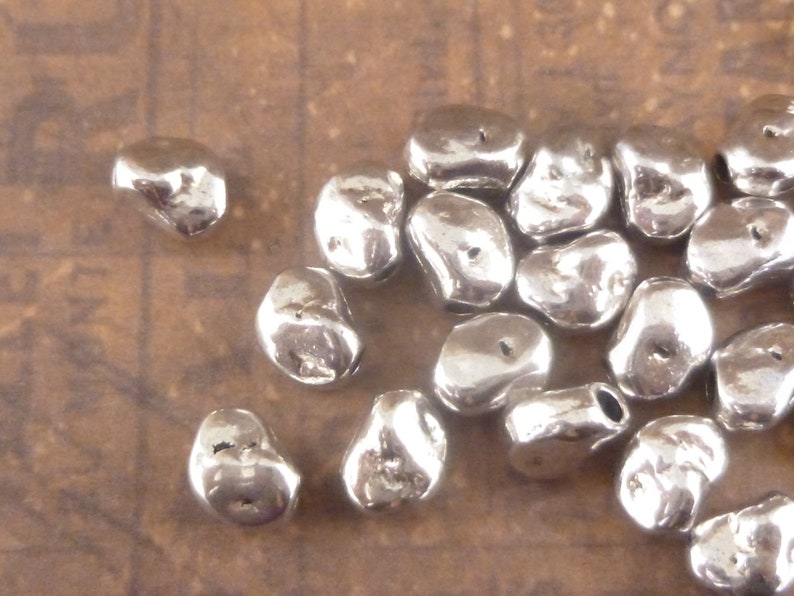 May include: A group of silver-colored metal beads, each with a unique irregular shape and a small hole for stringing.