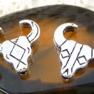 May include: Two silver-colored bull skull charms with detailed horns and facial features. Each charm has a unique geometric design on the forehead. The charms are displayed on a brown and black patterned surface.