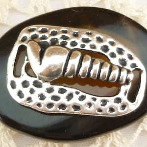 May include: A black and brown agate oval button with a silver metal design of a stylized horseshoe with a spiral inside. The button has a textured surface.