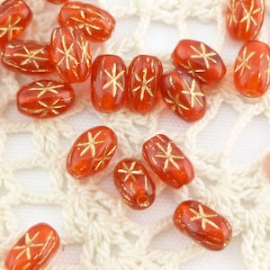 Könnte beinhalten: Eine Sammlung ovaler Glasperlen mit roter Basis und goldenen Sternmotiven. Die Perlen sind auf einer weißen Häkelunterlage verstreut.
