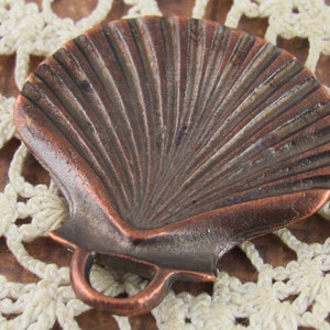 May include: A copper-colored metal seashell charm with a detailed, textured surface. The charm has a loop at the bottom for attaching to a necklace or bracelet.