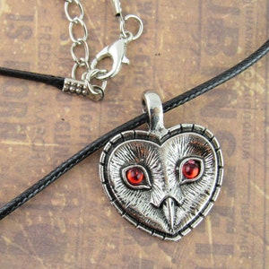 Peut inclure: Un collier avec un pendentif en forme de c&oelig;ur argent&eacute; repr&eacute;sentant un hibou d&eacute;taill&eacute; avec des yeux en pierres pr&eacute;cieuses rouges. Le collier est sur un cordon noir.