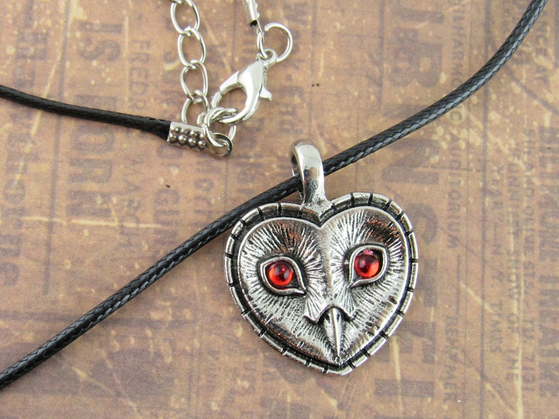 Peut inclure: Un collier avec un pendentif en forme de c&oelig;ur argent&eacute; repr&eacute;sentant un hibou d&eacute;taill&eacute; avec des yeux en pierres pr&eacute;cieuses rouges. Le collier est sur un cordon noir.