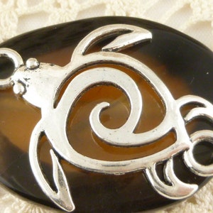 May include: A brown and black oval-shaped pendant with a silver turtle design. The turtle is outlined in silver and has a spiral design in the center.