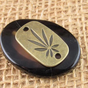 May include: A smooth, dark brown oval stone cabochon layered with a rectangular, antique brass-toned metal tag. The metal tag features a centered, engraved cannabis leaf design and two circular holes, one at the top and one at the bottom edge.