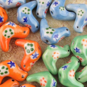 May include: A collection of ceramic beads in orange, blue, and green. Each bead has a unique shape with a hole for stringing. The beads are decorated with white polka dots and blue star-shaped floral designs.