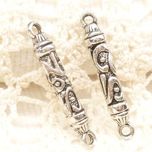 May include: Two silver-toned metal pendant charms with intricate embossed designs. Each charm is a long, cylindrical shape with a loop at each end for attaching to jewelry. The charms have a vintage or antique appearance.