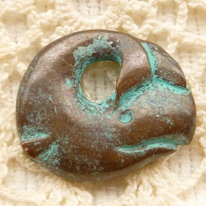 May include: A small, round, bronze pendant with a hole in the centre. The pendant has a green patina and is shaped like a fish.