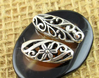 May include: Two silver-colored, filigree oval beads with intricate openwork designs. One bead features a floral center. The beads rest on a dark, polished, oval-shaped surface, set against a textured, tan background.
