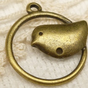 Puede incluir: Un amuleto de metal de color bronce que presenta un pequeño pájaro dentro de un círculo. El amuleto tiene un agujero en la parte superior para sujetarlo a un collar o pulsera.