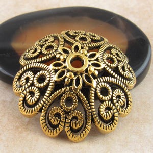 May include: A gold-coloured filigree bead cap with an intricate floral design. The bead cap has a central flower shape and is surrounded by swirling, openwork patterns. The bead cap is approximately 3.8 cm in diameter.