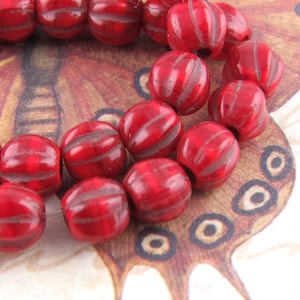 May include: Close-up of a strand of red, round beads with a striped design. The beads are arranged on a decorative background featuring a butterfly illustration in shades of brown and tan. The beads have a glossy finish.