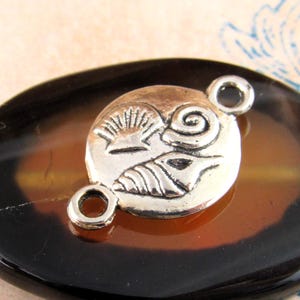 May include: A silver-toned metal connector charm featuring a circular design with embossed seashell motifs. The charm includes two small circular loops on opposite sides for attachment, showcasing a detailed relief of various sea shells.