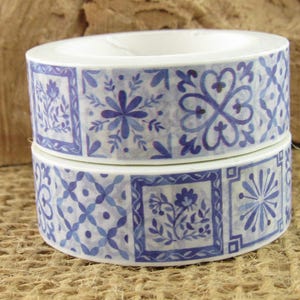 May include: Two rolls of adhesive tape stacked vertically, featuring a repeating pattern of blue and white square tiles. The designs include floral motifs, geometric star shapes, and intricate lattice patterns printed on a smooth surface.