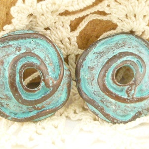 May include: Two round, weathered metal pendants with a spiral design. The pendants have a turquoise patina over a bronze base, with a hole in the center of the spiral. The background is a cream-colored crocheted doily.