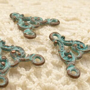 May include: Three ornate metal triangle-shaped charms with a turquoise patina finish. The charms have a decorative scroll design and two open loops.
