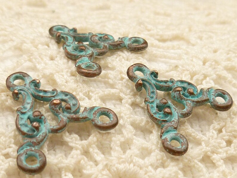 May include: Three ornate metal triangle-shaped charms with a turquoise patina finish. The charms have a decorative scroll design and two open loops.