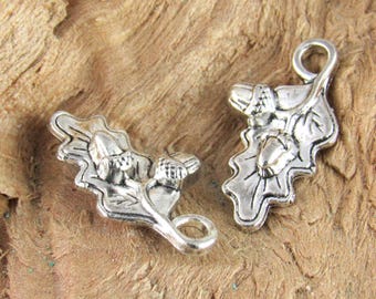 May include: Two silver-colored acorn and oak leaf charms. Each charm features detailed leaf and acorn designs, with a small loop for attaching to jewelry or other crafts. The charms are set against a textured, brown background.