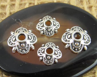May include: Four silver-colored metal bead caps with a floral design. Each cap has a central hole and decorative details. The caps are arranged on a dark, oval-shaped surface.