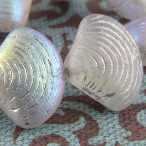 Puede incluir: Una colección de cuentas de vidrio blanco iridiscente con forma de conchas marinas. Las cuentas tienen una superficie texturizada y un brillo sutil.