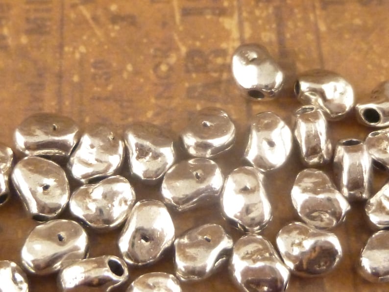 May include: A close-up of a pile of silver-colored metal beads. The beads are irregularly shaped and have a slightly rough texture. They are perfect for jewelry making or other craft projects.