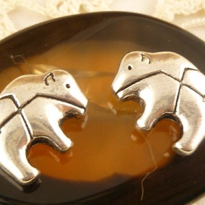 May include: A pair of silver-toned bear-shaped earrings. Each bear has a detailed design with an arrow across its body. The earrings are displayed on a brown and amber-colored oval stone.
