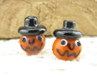 Può includere: Due perline di zucca in vetro arancione con cappelli neri e volti dipinti. Le perline hanno occhi bianchi e bocche nere.