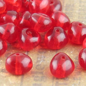 Peut inclure: Un assortiment de perles rouges translucides en forme de goutte d'eau. Chaque perle est percée d'un trou pour l'enfilage et présente une finition brillante. Les perles sont dispersées sur une surface texturée.