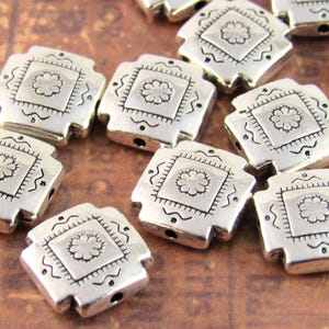 May include: A collection of square silver-toned metal beads, each featuring a central embossed flower design within a square border, accented by small dots and wavy lines along the edges, with a hole visible on the side of each individual bead.