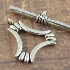 May include: Silver tone toggle clasp with a decorative, ornate design. The clasp is made of two pieces: a bar and a frame. The bar is straight and has a rounded end. The frame is shaped like a square with rounded corners. The frame has a decorative design of raised lines and circles.