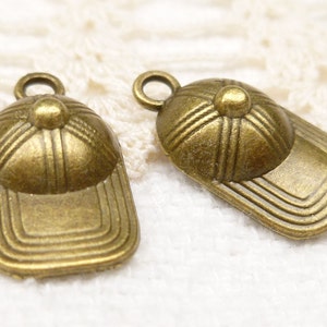 Può includere: Due ciondoli in metallo color oro antico a forma di cappellini da baseball. I ciondoli hanno un design dettagliato e un anello per attaccarli a una collana o un braccialetto.
