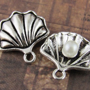May include: Two silver-toned metal seashell charms with a pearl accent. The seashells are open and have a detailed design. The charms have a loop for attaching to jewelry.